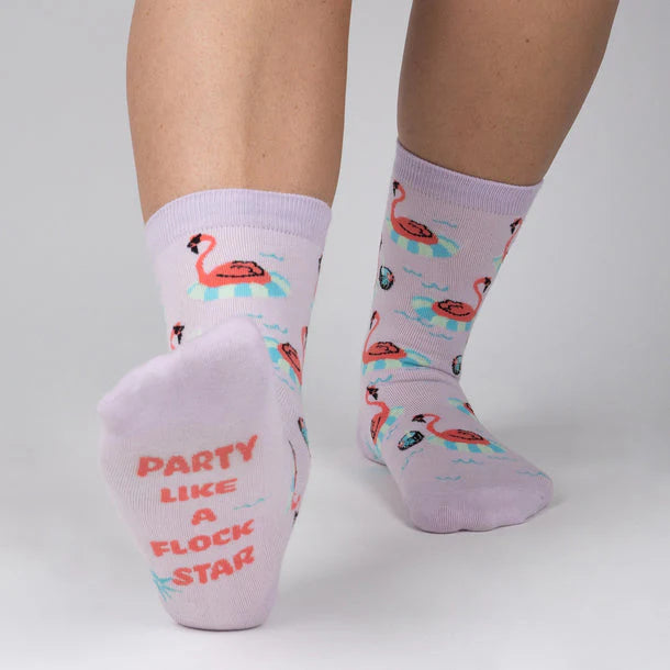 Pastel purple socks with flamingos wearing sunglasses floating around in pool rings and sole of sock ahs pink text "party like a flock star".