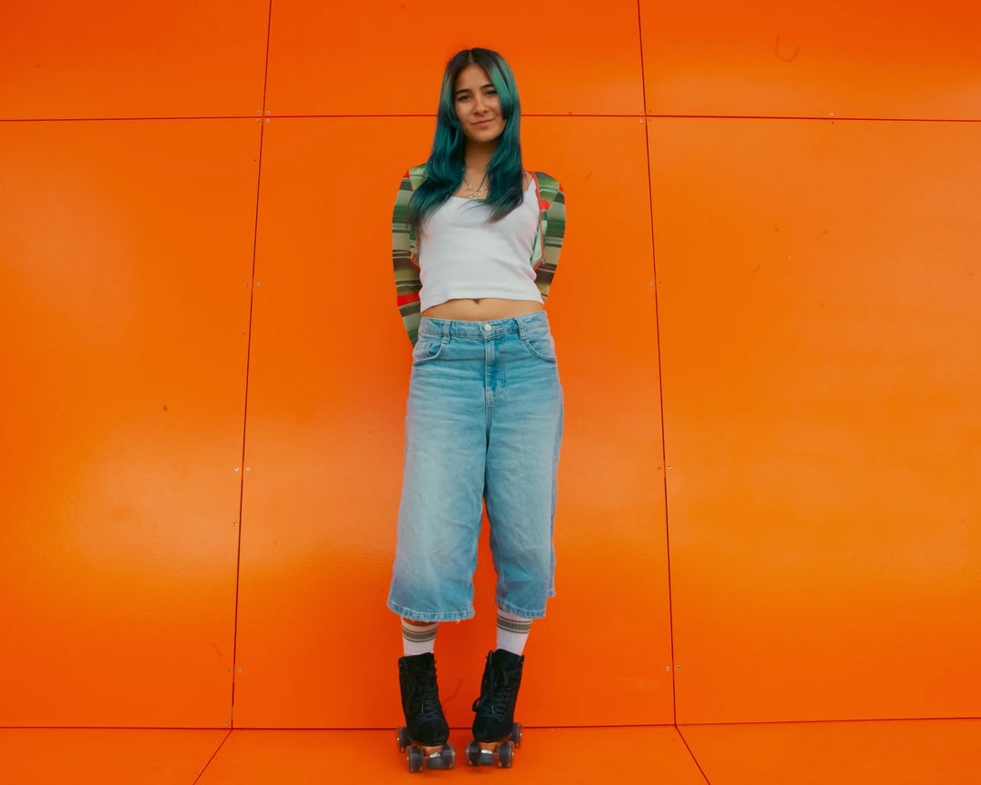 Rib stands against an orange wall wearing a white and green top, light denim jorts, white socks and Chuffed Skates Wanderer Roller Skates in Black - a sleek looking suede skate.