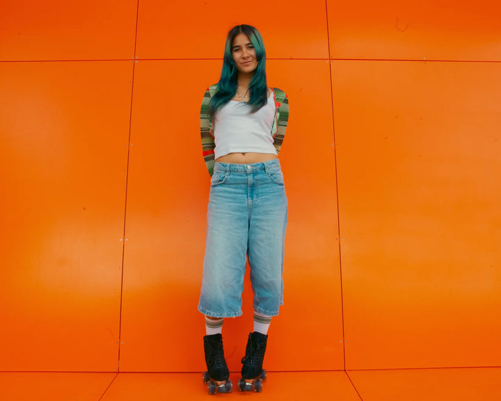 Rib stands against an orange wall wearing a white and green top, light denim jorts, white socks and Chuffed Skates Wanderer Roller Skates in Black - a sleek looking suede skate.