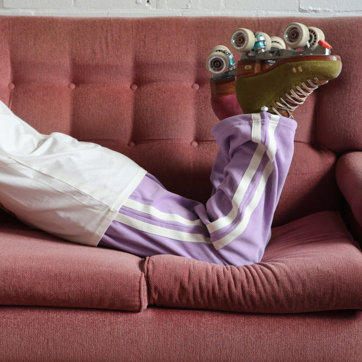 Stacey wears the purple wide leg stripe track pants from the RollerFit Shop with her roller skates as she lays on a pink sofa.