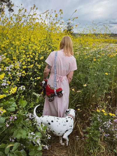 Person wearing beige gingham dress in a flower field carrying red roller skates with dalmatian strap and Dalmatian dog following behind.
