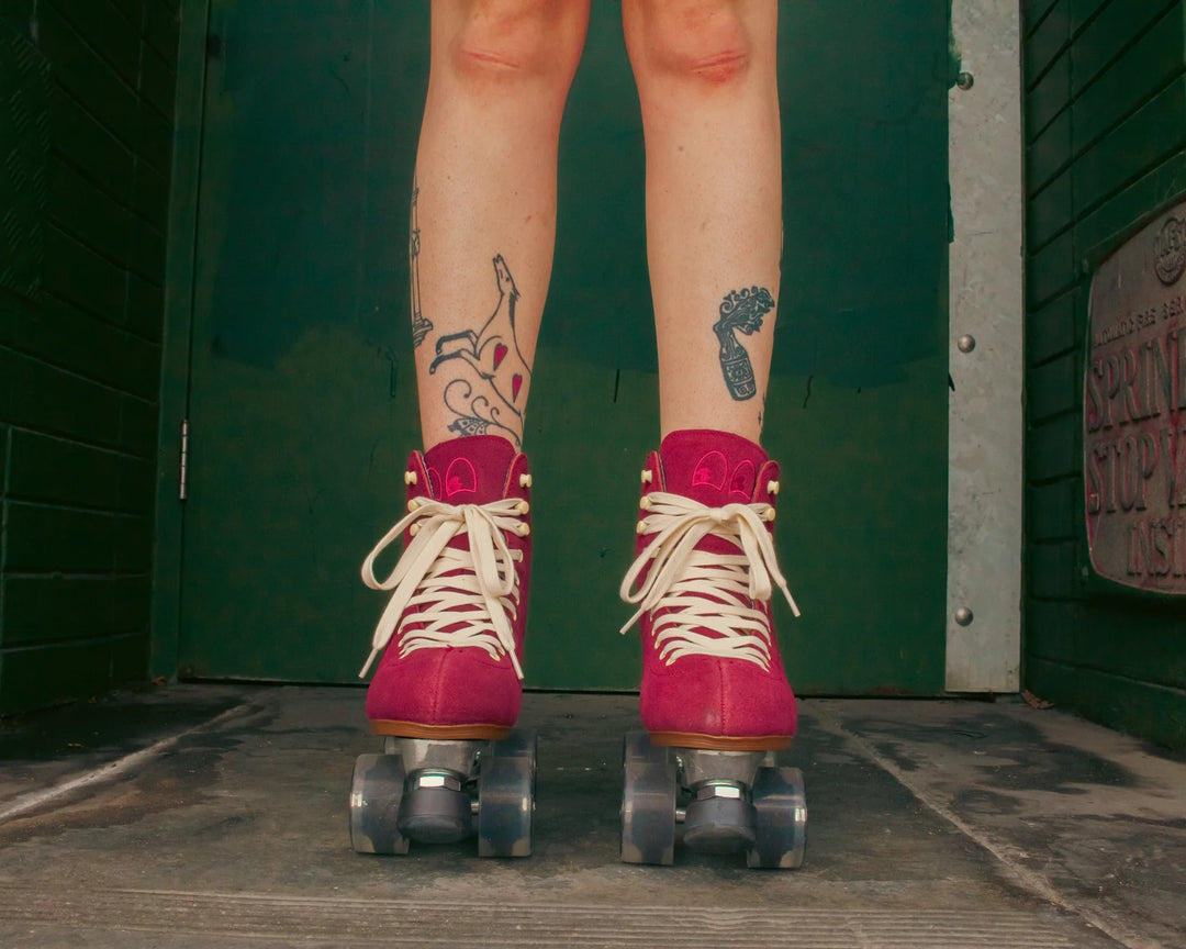 Roller skater with tattooed legs wears Chuffed Skates Wanderer roller skates in Burgundy against a dark green wall.