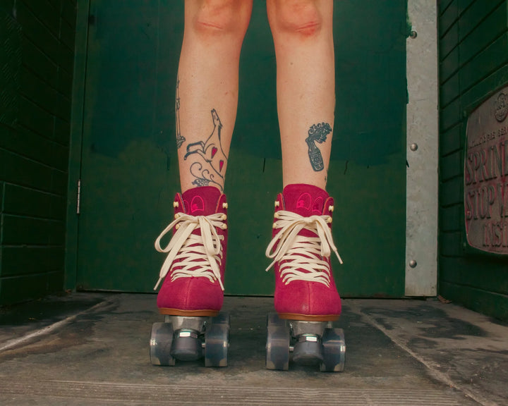 Roller skater with tattooed legs wears Chuffed Skates Wanderer roller skates in Burgundy against a dark green wall.