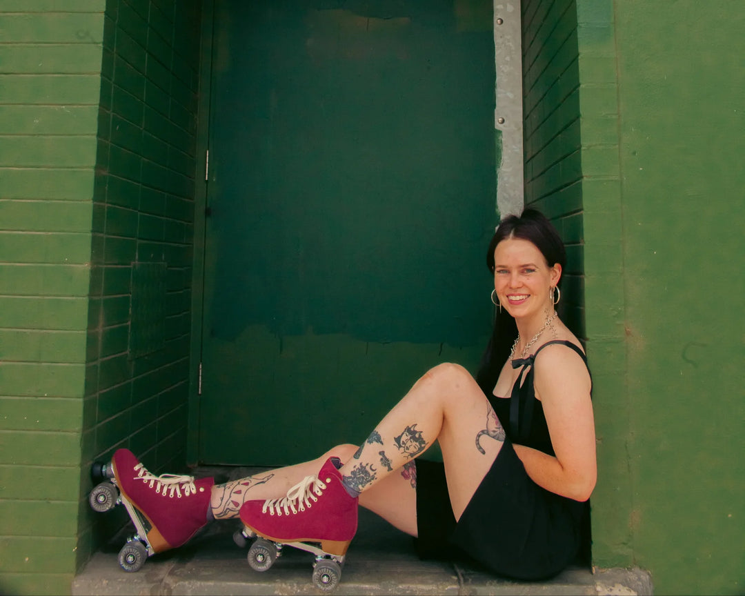 Roller skater with tattooed legs wears black short jumpsuit with Chuffed Skates Wanderer roller skates in Burgundy as they sit against a dark green wall.