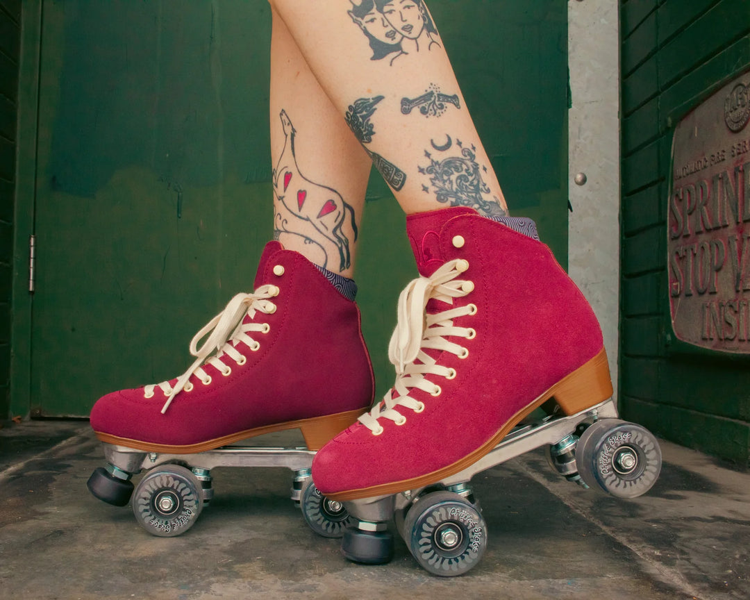 Roller skater with tattooed legs wears Chuffed Skates Wanderer roller skates in Burgundy against a dark green wall.