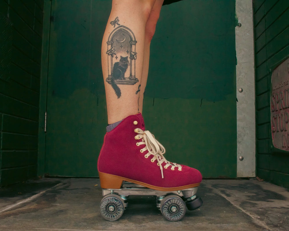 Roller skater with tattooed legs wears Chuffed Skates Wanderer roller skates in Burgundy against a dark green wall.