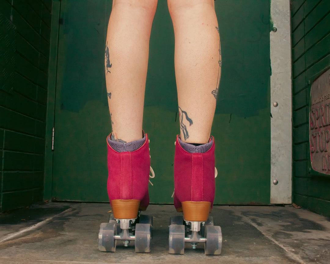 Roller skater with tattooed legs wears Chuffed Skates Wanderer roller skates in Burgundy against a dark green wall.