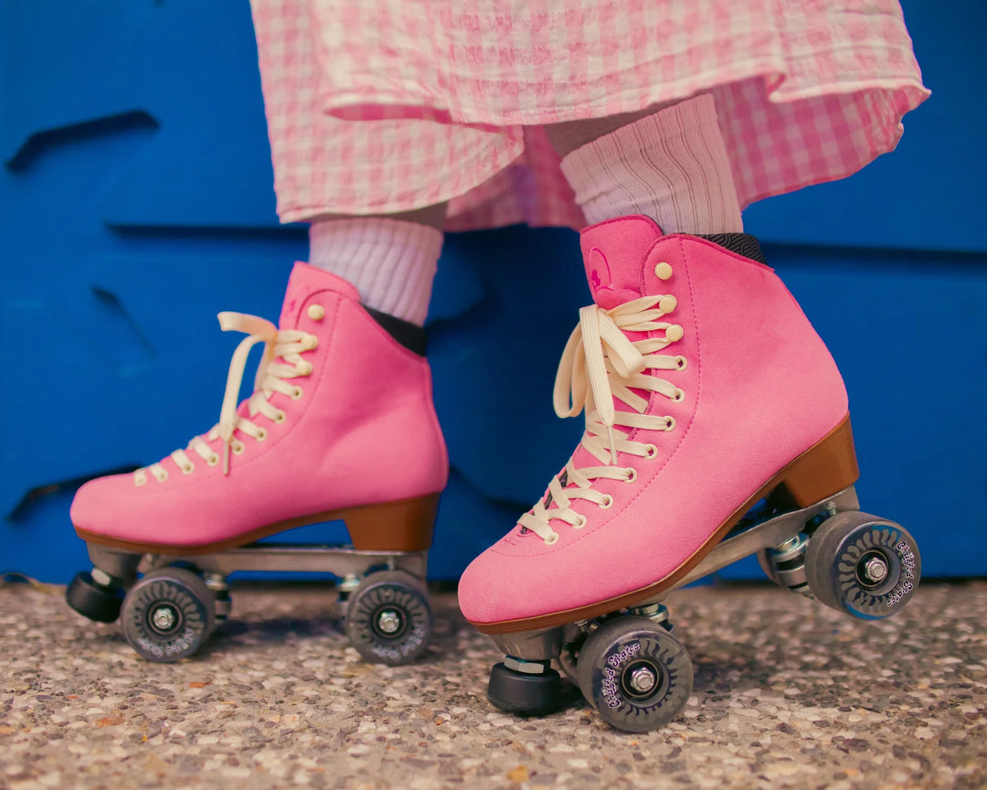 Roller skater standing side on against a blue wall pictured from below the knee wearing a pink and white gingham skirt with Chuffed Skates Wanderer Vegan Pink roller skates.