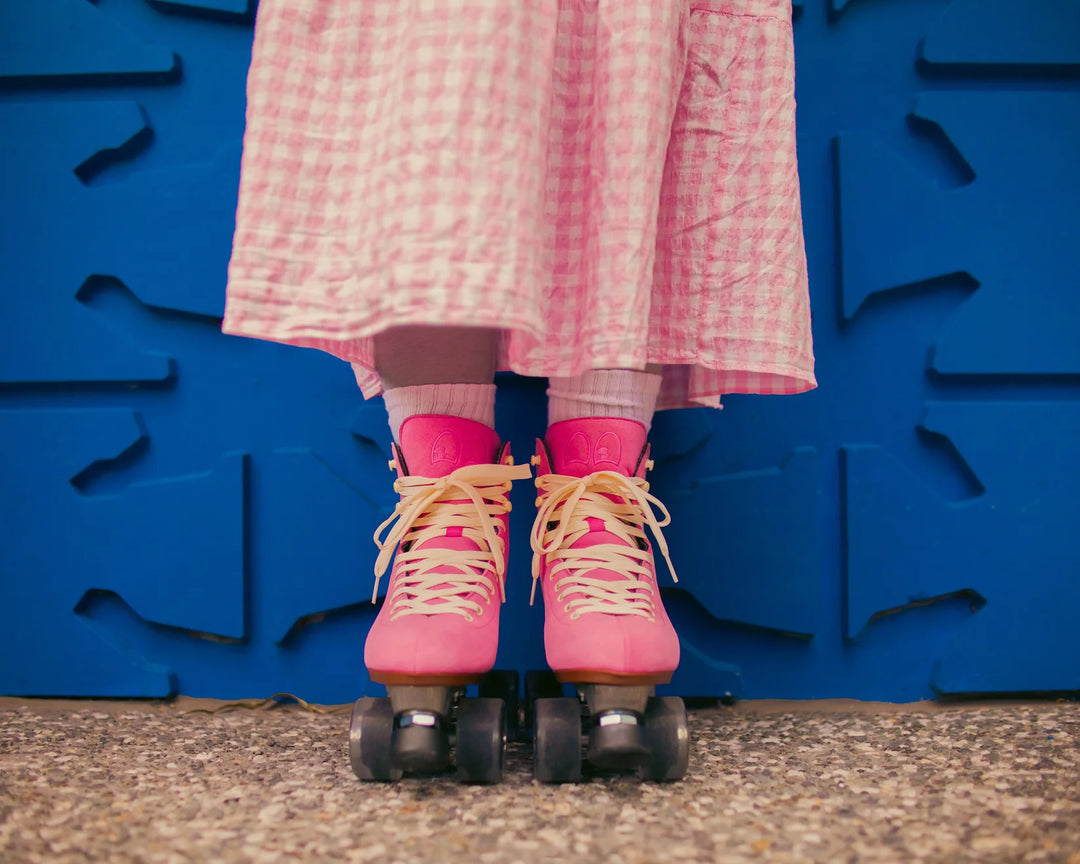 Roller skater standing front on against a blue wall pictured from below the knee wearing a pink and white gingham skirt with Chuffed Skates Wanderer Vegan Pink roller skates.