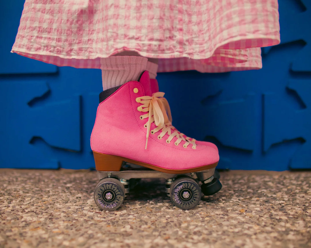 Roller skater standing side on against a blue wall pictured from below the knee wearing a pink and white gingham skirt with Chuffed Skates Wanderer Vegan Pink roller skates.