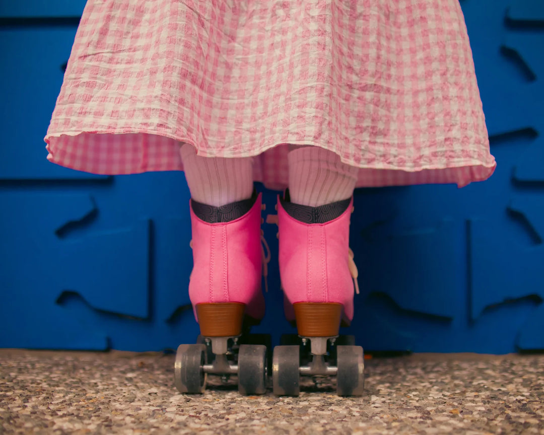 Roller skater standing  against a blue wall pictured from below the knee wearing a pink and white gingham skirt with Chuffed Skates Wanderer Vegan Pink roller skates.