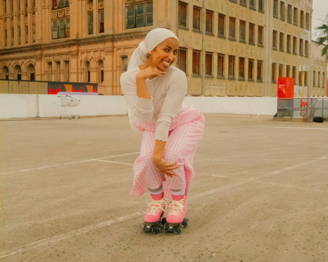 Zulfiye wears a white hijab and long sleeve top with a pink gingham skirt and Vegan Pink Chuffed Skates Wanderers as she roller skates through a carpark with a big smile on her face.