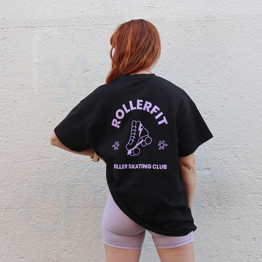 Person wearing a black oversized t-shirt with 'Rollerfit' and 'Roller Skating Club' text against a light gray wall.