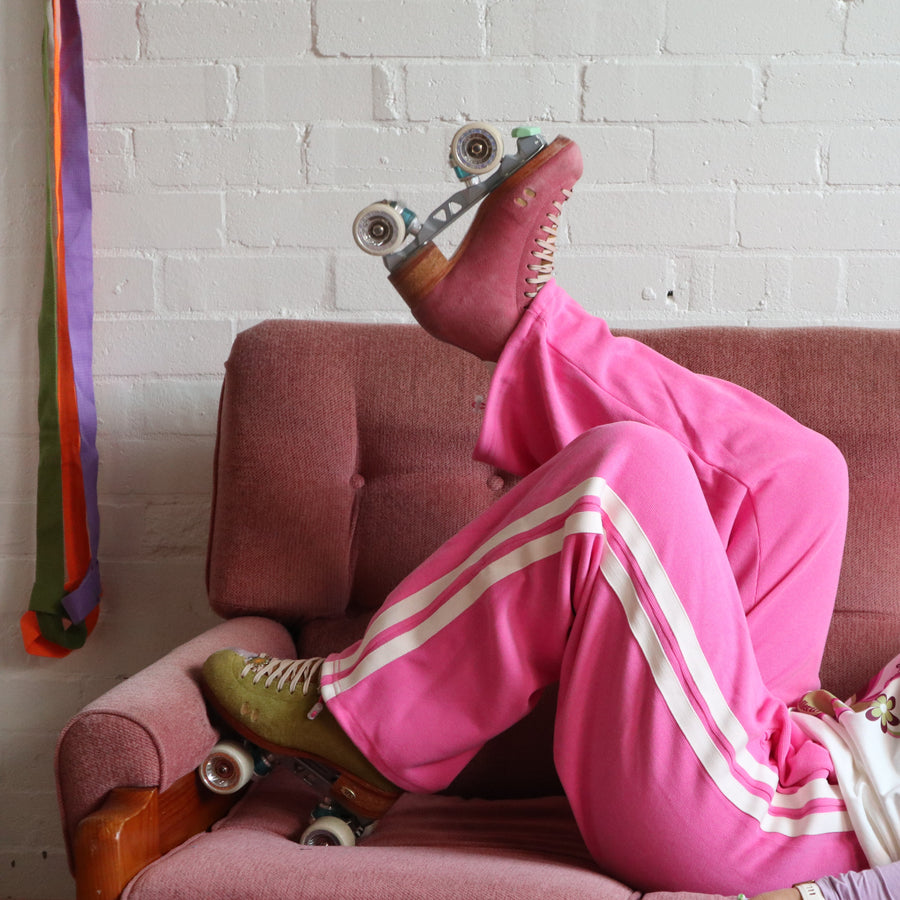 Stacey wears the pink wide leg stripe track pants from the RollerFit Shop with her roller skates as she lays on a pink sofa.