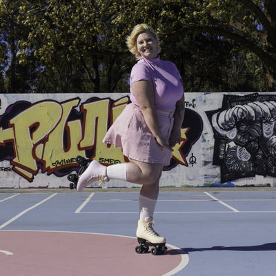 Emily is skating on a basketball court wearing a lilac t-shirt and pale pink skirt and Sunset Cruiser roller skates.