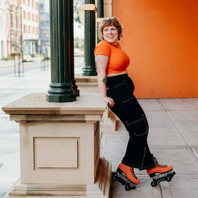 Sophie wears black long pants and an orange crop top leaning against a sandstone pillar with her Chuffed Skates Wild Thing Pro Boots, orange suede with black laces and soles, yellow/black tiger print and floral lining.