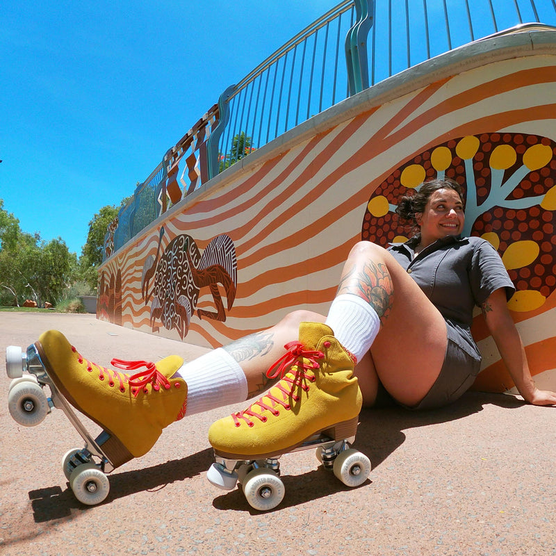 Ashley sits against an Indigenous mural wearing the Birak mustard yellow and coral pink roller skates.