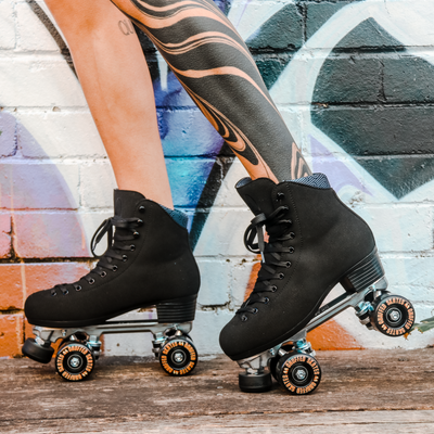 Ash with tattooed legs wears Chuffed Skates Wanderer roller skates in Vegan Black against a graffiti brick wall.