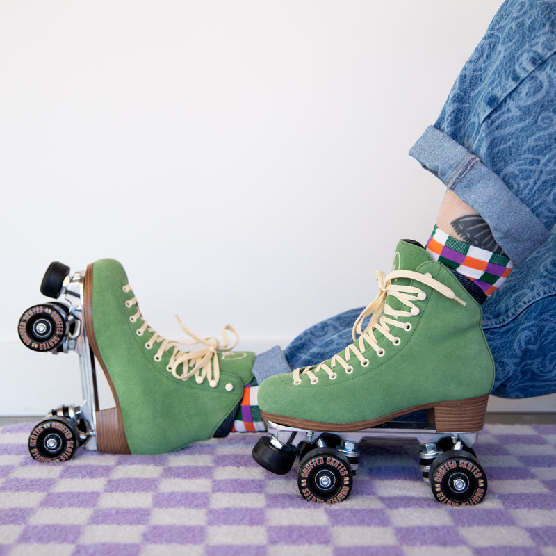 Sophie sits on a purple and white check rug and is wearing Chuffed Skates Wanderer roller skates in Olive Green with cream laces, eyelets and logo embroidery on the tongue, black wheels and toe stops.