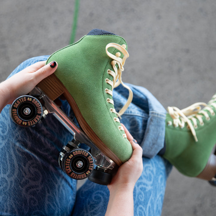 Sophie is wearing blue jeans and is holding Chuffed Skates Wanderer roller skates in Olive Green with cream laces, eyelets and logo embroidery on the tongue, black wheels and toe stops.