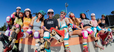 10 roller skaters hang their legs into a skate bowl, wearing Moxi x Triple 8 helmets and Moxi x 187 six pack pads.
